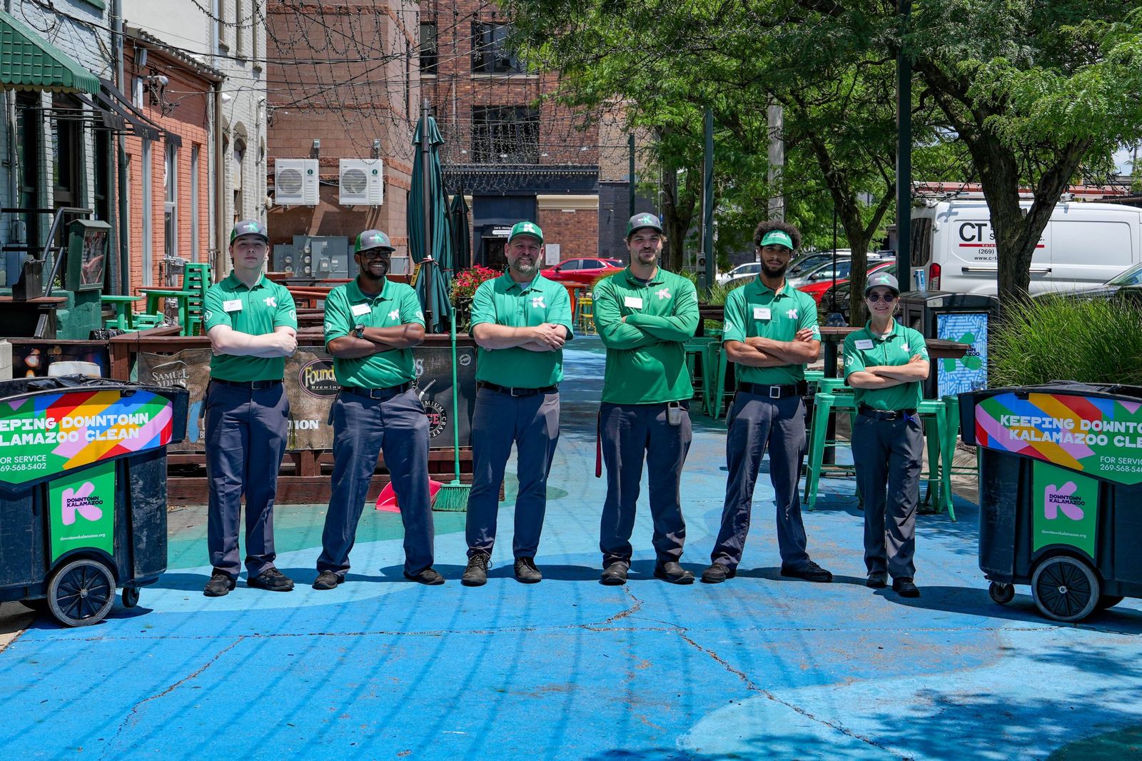 Six Downtown Ambassadors in green Downtown Kalamazoo shirts and caps stand arms crossed in a painted blue alley, between two branded cleaning carts.